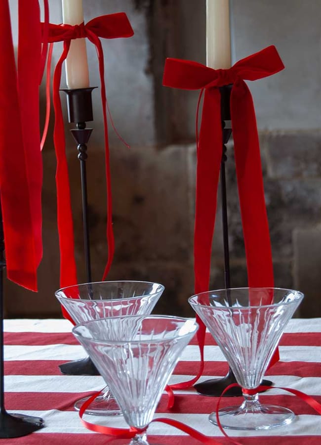 Three empty martini glasses sit on a red and white striped tablecloth next to three tall black candlesticks with white candles, each tied with a large red ribbon—creating a festive Christmas tablescape.
