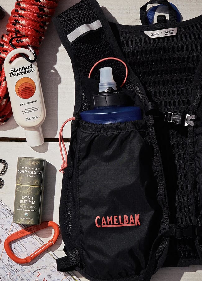 A CamelBak hydration pack, sun cream, soap & salve, carabiner, and orange rope are arranged on a surface with a partially visible map in the background—perfect kit to pair with the latest activewear for your next adventure. The CamelBak logo is prominent on the pack.