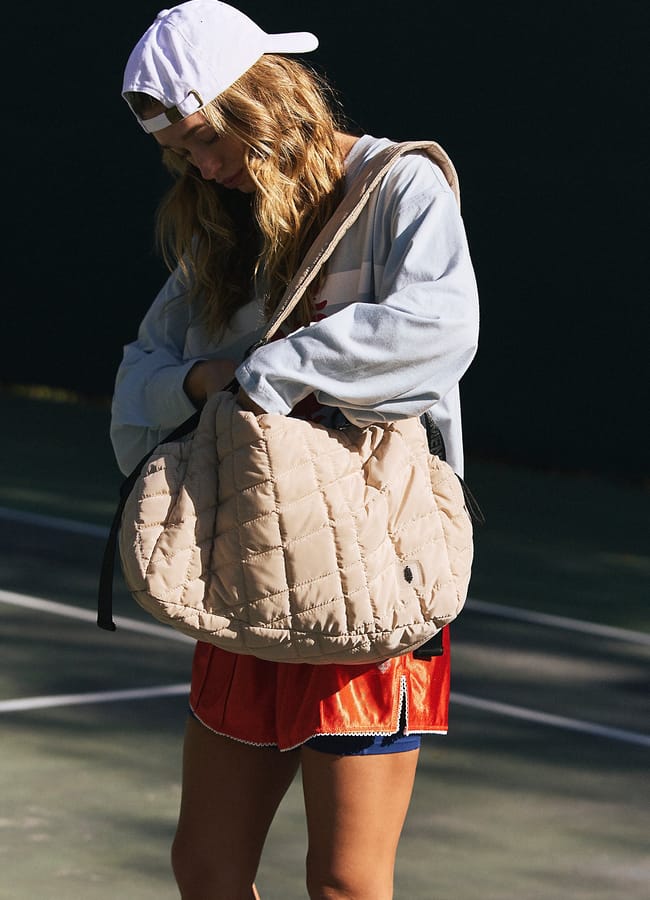 A woman wearing a white cap, light jacket, and red sports shorts stands outdoors on a sports court, looking into a large, quilted beige bag slung over her shoulder—showcasing the latest activewear trends for the best women's activewear look.
