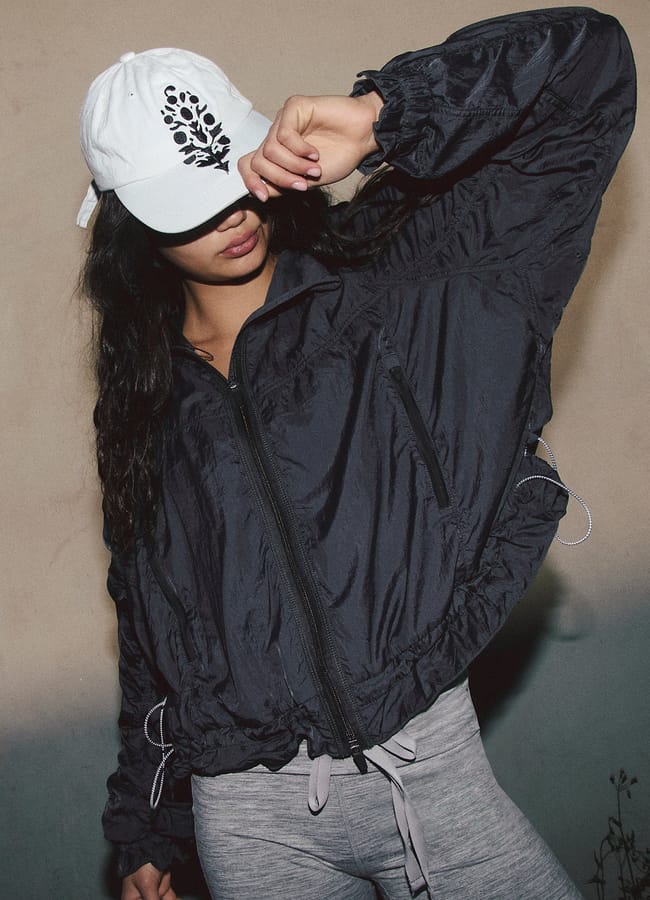 A person in the latest activewear—a white baseball cap with a black design, a black zip-up jacket, and light grey leggings—poses with one arm across their face, standing against a plain wall.