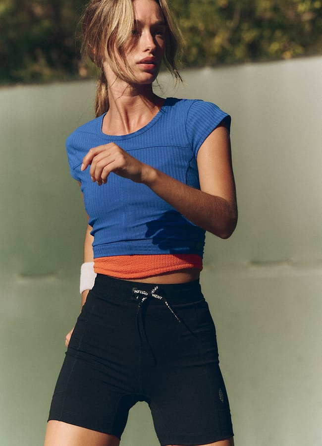 A woman stands outdoors in the latest activewear, wearing a blue fitted top, black high-waisted shorts, and an orange waistband. Her light brown hair is tied back as she looks to the side in natural sunlight against a blurred green background.