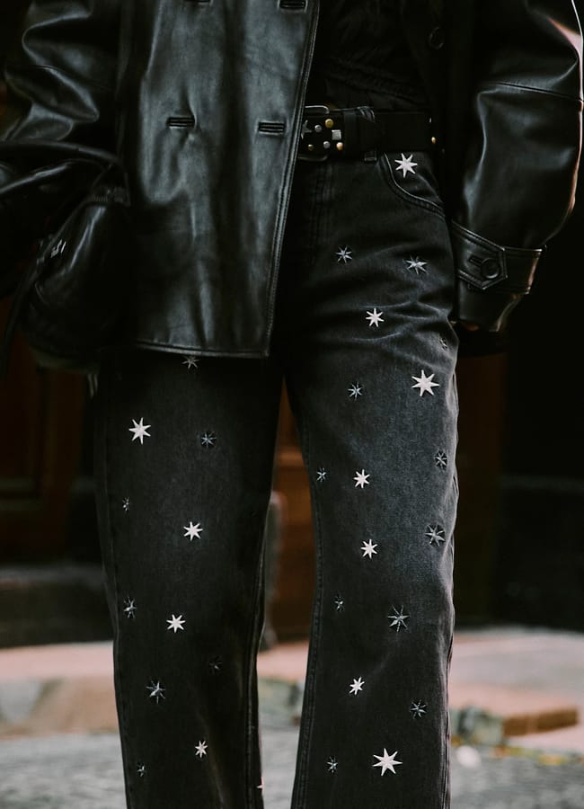 A person wears a black leather jacket and embellished jeans decorated with white star patterns, standing outdoors on a stone pavement.