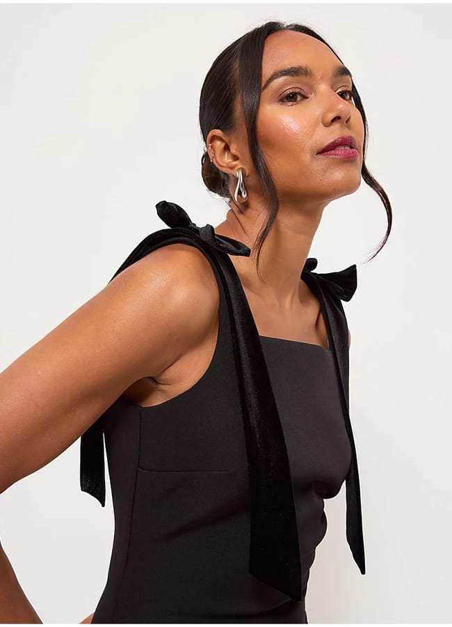 A woman wearing a sleeveless black dress with large velvet bows on the shoulders showcases chic bow fashion as she poses against a plain white background, her hair tied back and gaze confidently to the side.
