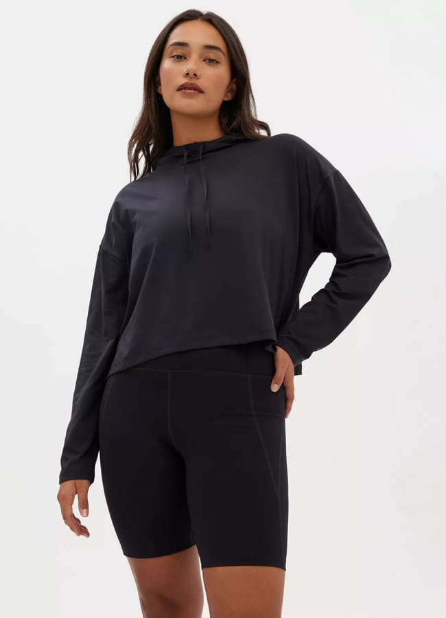A woman stands against a plain light background wearing the latest activewear: a loose-fitting black long-sleeve hoodie and fitted black cycling shorts, with one hand resting on her hip.