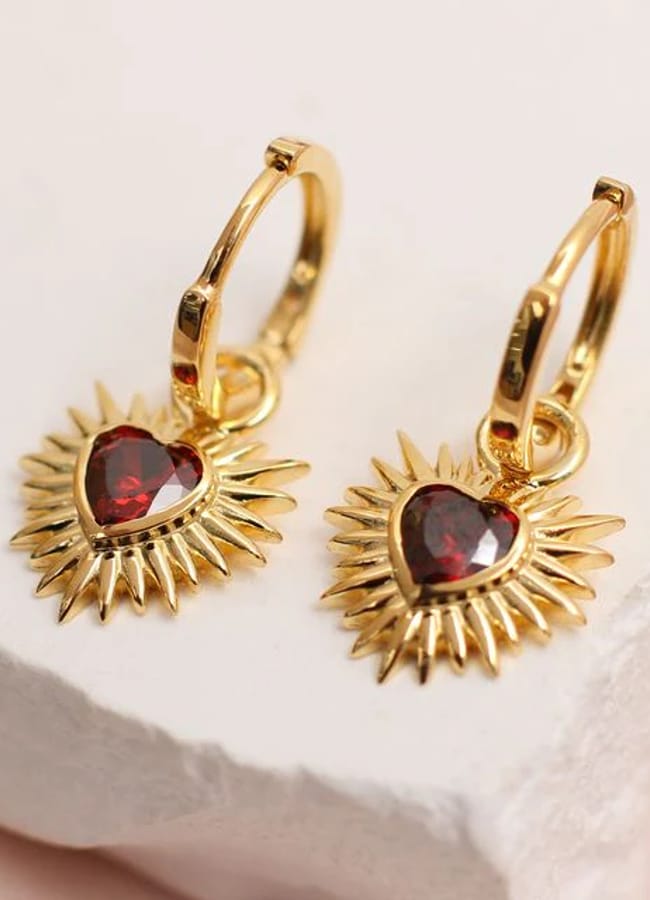 A pair of gold hoop earrings featuring heart-shaped red gemstones set in the centre of sunburst designs, perfect for adding festive sparkle to your Christmas tablescape, displayed on a white stone surface.