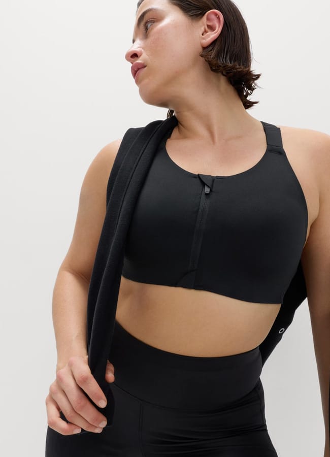 A person wearing the latest activewear—a black sports bra and leggings—looks to the side, holding a black jacket over one shoulder against a plain light background.
