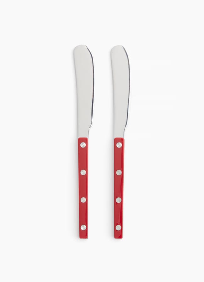 Two butter knives with shiny silver blades and glossy red handles, each handle featuring four silver rivets, are placed side by side on a white background—a subtle nod to Unexpected Red Theory in interior design.