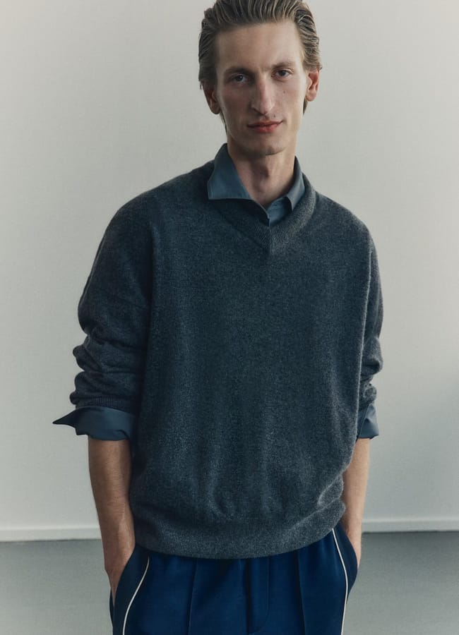 A person with swept-back blond hair stands against a plain background, wearing a grey V-neck men's jumper over a blue collared shirt and dark blue trousers, hands casually in their pockets.