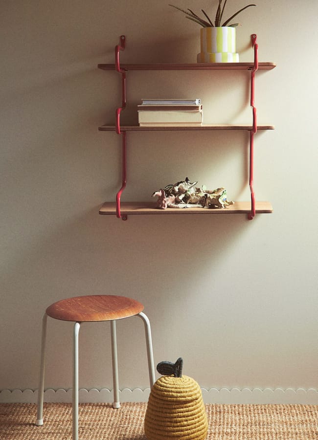 A minimalist wall shelf with red brackets, inspired by the Unexpected Red Theory in interior design, holds a potted plant, books, and small animal figurines above a woven basket and a simple stool on a textured carpet.
