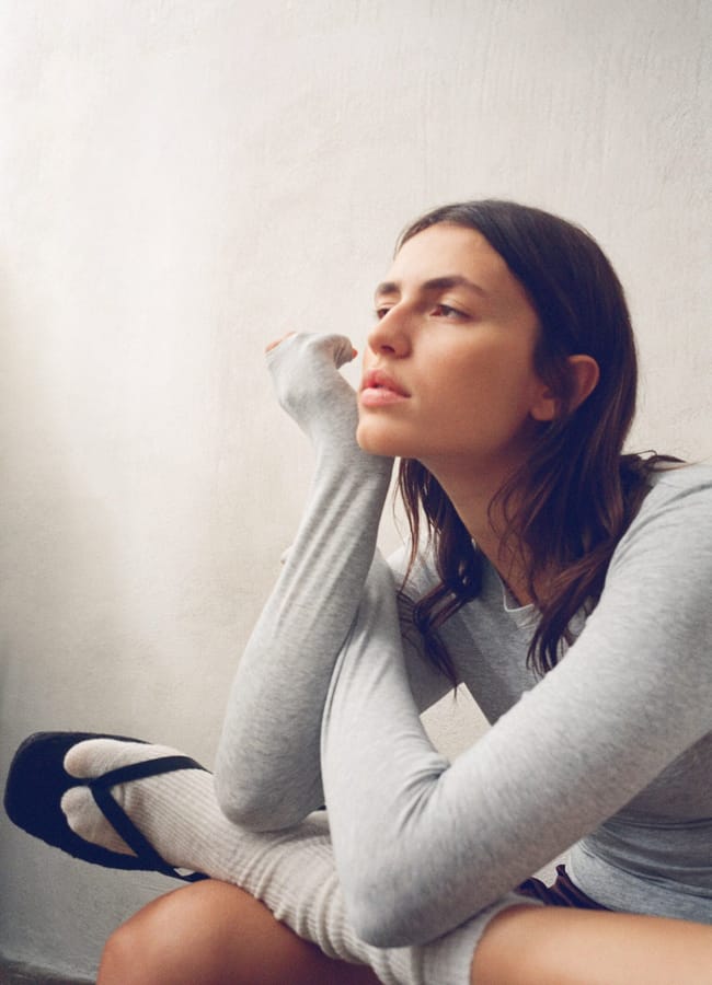 A young woman with long brown hair sits with her chin resting on her hand, wearing the latest sportswear: a light grey long-sleeve shirt, white socks, and black sandals, gazing thoughtfully into the distance against a plain wall.
