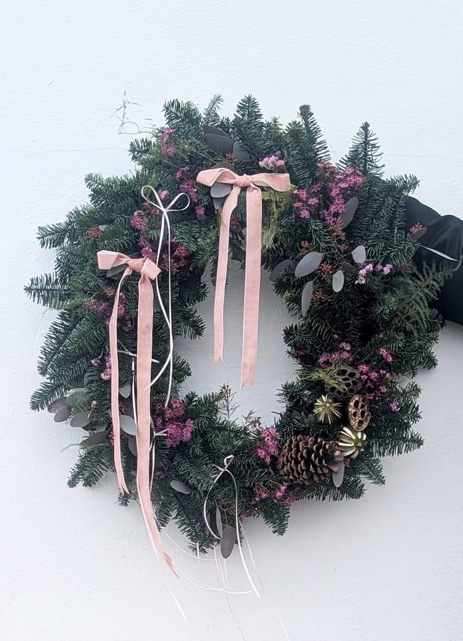 A festive green wreath decorated with pink bows, purple flowers, pine cones, and gold accents is held against a white wall—perfect inspiration for your Christmas tablescape.
