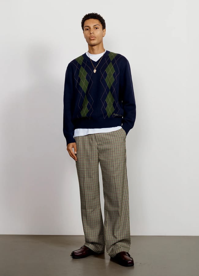 A person stands against a plain white wall, wearing a navy blue men's jumper with a green argyle pattern, a white T-shirt underneath, loose checked trousers, brown shoes, and a necklace.