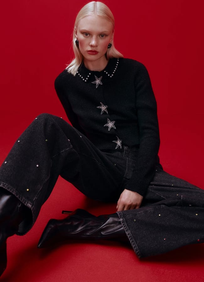 A woman with straight blonde hair sits against a red background, wearing a black jumper with star-shaped buttons, black embellished jeans with small coloured dots, and black knee-high boots. She has a serious expression.