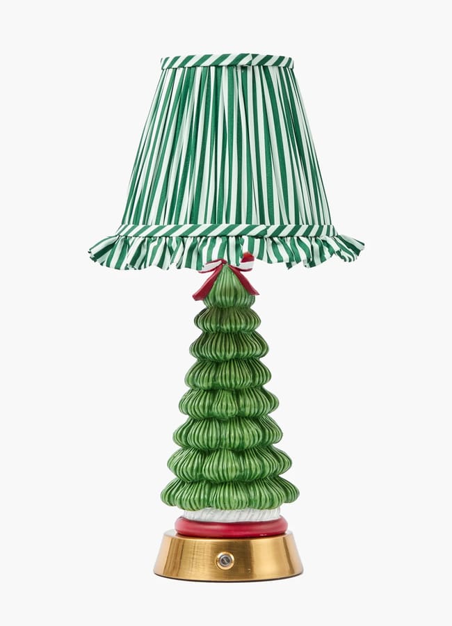 A festive table lamp with a green ceramic base shaped like a Christmas tree, perfect for elevating your Christmas tablescape. It features a red bow, a green-and-white striped ruffled lampshade, and rests on a gold circular base.