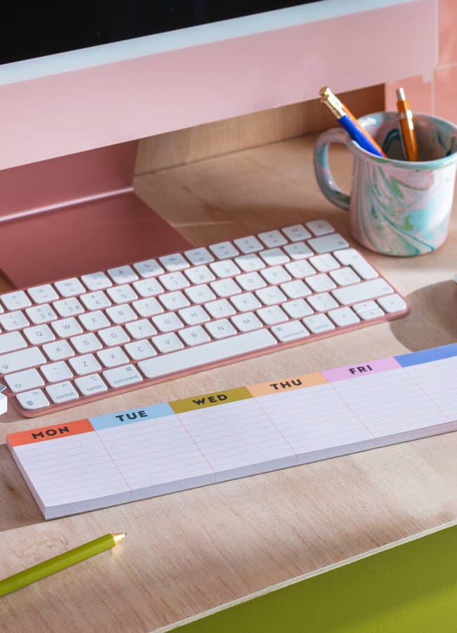 A tidy desk with a pink computer, white keyboard, mouse, 2026 diaries, a weekly planner pad, a multicoloured mug holding pencils, and a green pencil on a light wood surface.