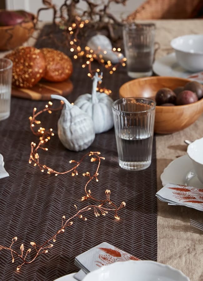A Thanksgiving table setting with white crockery, glasses of water, serviettes with leaf prints, a brown table runner, bread rolls, a wooden bowl of plums, and decorative fairy lights with white pumpkins creates a cosy Christmas tablescape.