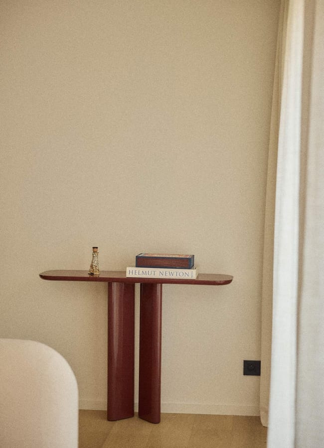 A minimalist scene inspired by Unexpected Red Theory: a small red console table stands against a beige wall, holding a decorative bottle and Helmut Newton book—perfect for 2026 interior design trends—beside a white curtain and light wood floor.