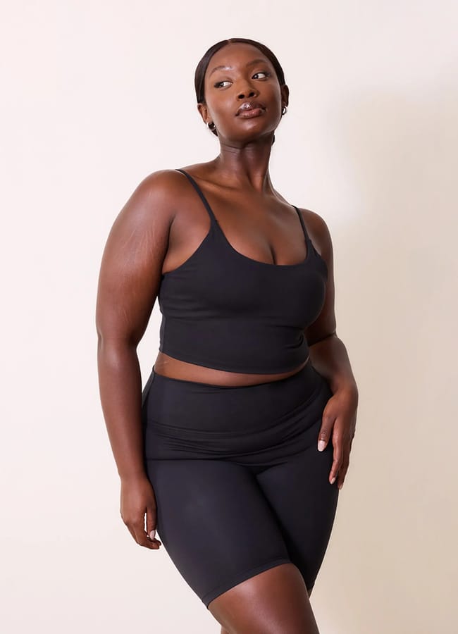 A woman wearing the latest activewear—a black vest top and high-waisted black cycling shorts—stands against a plain light background, looking confidently to the side with one hand resting on her hip.