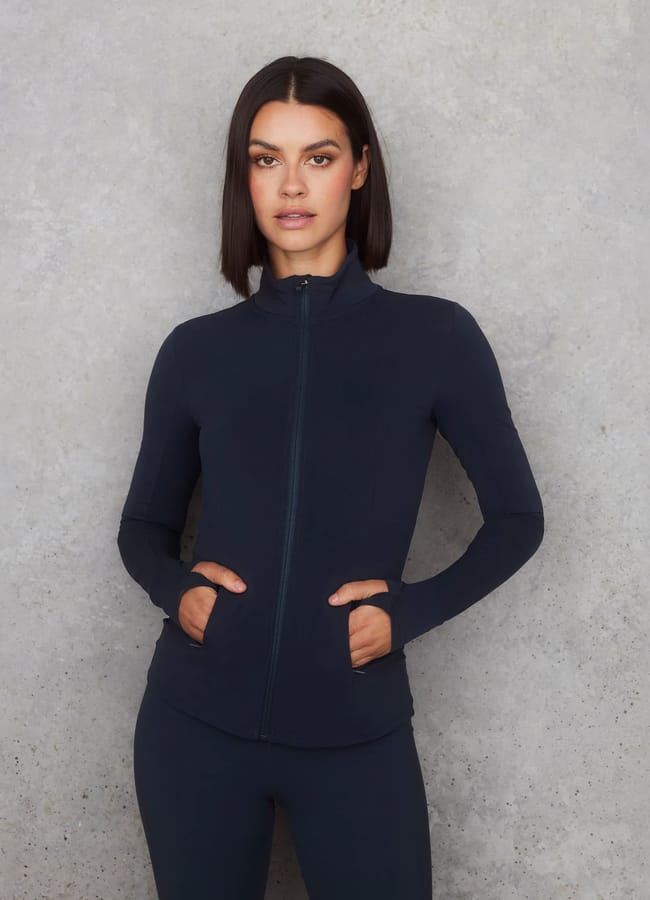 A woman with straight dark hair stands against a light grey textured wall, wearing the latest activewear—a fitted, long-sleeve navy athletic zip-up jacket and matching leggings. Her hands are in the jacket pockets.