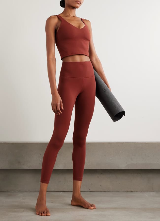 A person wearing the best women’s activewear—a matching rust-coloured sports bra and high-waisted leggings—stands barefoot on a smooth floor, holding a rolled-up grey yoga mat by their side against a neutral background.
