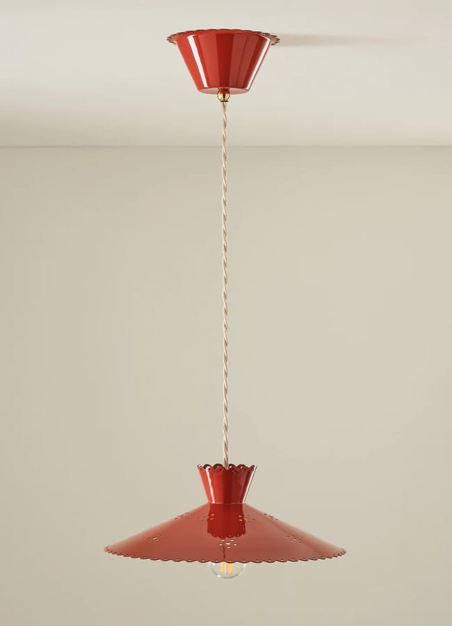 A red pendant light with a scalloped-edged shade hangs from the ceiling on a twisted cord, embodying the Unexpected Red Theory—an eye-catching accent for any interior design in 2026 against a plain beige wall.
