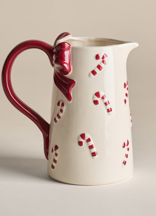 A cream-coloured ceramic jug with a red handle and bow, decorated with small red and white candy cane designs. Perfect for adding festive charm to your Christmas tablescape, the jug is set against a neutral background.