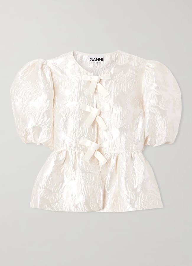 A short-sleeved, textured ivory blouse with puffed sleeves, three front ribbon bows for a touch of bow fashion, and a ruffled waist. The blouse features shiny, crinkled fabric and a round neckline with a visible GANNI label inside the collar.