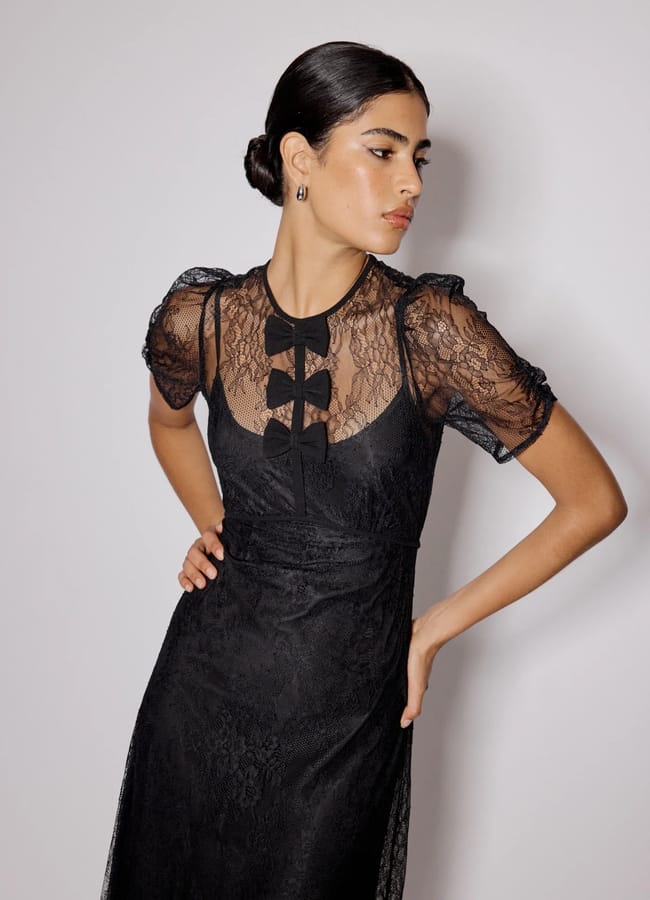 A woman wearing a sheer black lace dress with puffed sleeves and bow fashion accents on the bodice stands against a plain light background, looking to the side with one hand on her hip.
