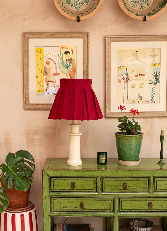 A green vintage-style chest of drawers styled with potted plants, candlesticks, and a white lamp with a red pleated shade illustrates Unexpected Red Theory—an interior design trend to watch in 2026. Framed art prints and decorative plates complete the look above.