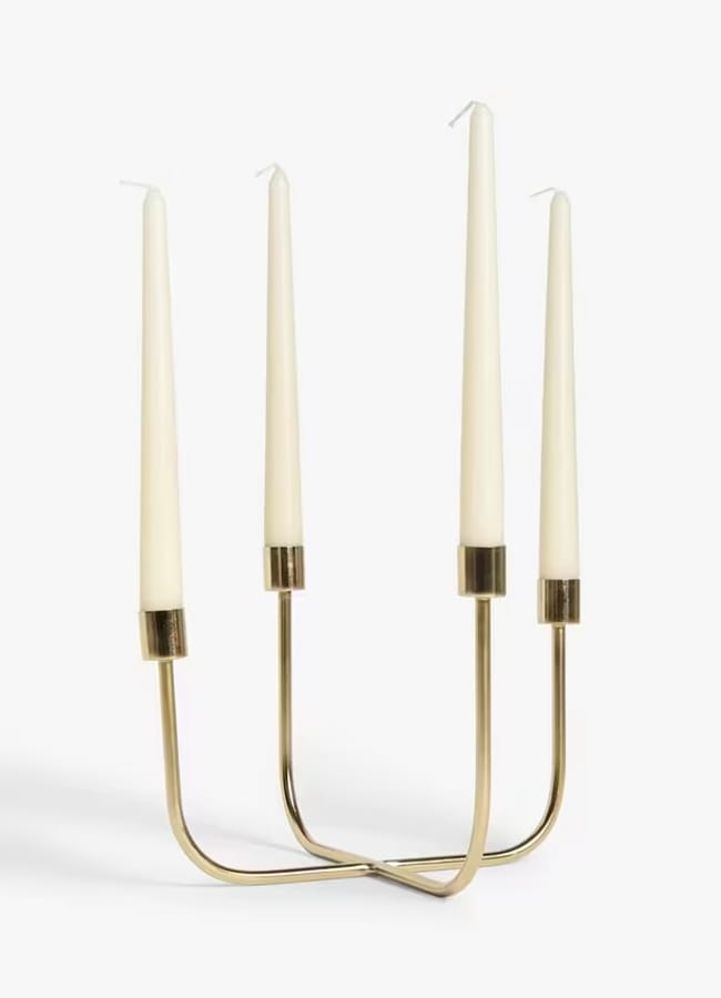 A modern gold metal candelabrum with four tall, white taper candles, each held at the top of gracefully curved stems, makes a stunning centrepiece for a Christmas tablescape against a plain white background.