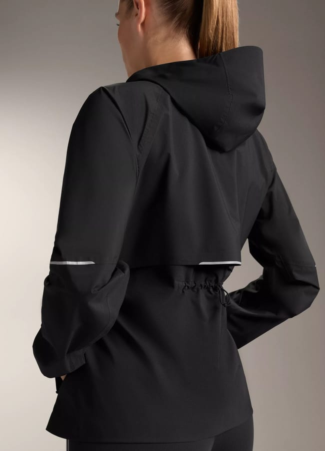 A woman with a ponytail is shown from the back, wearing the latest sportswear—a black hooded jacket with reflective strips on the sleeves and back—standing against a neutral background.