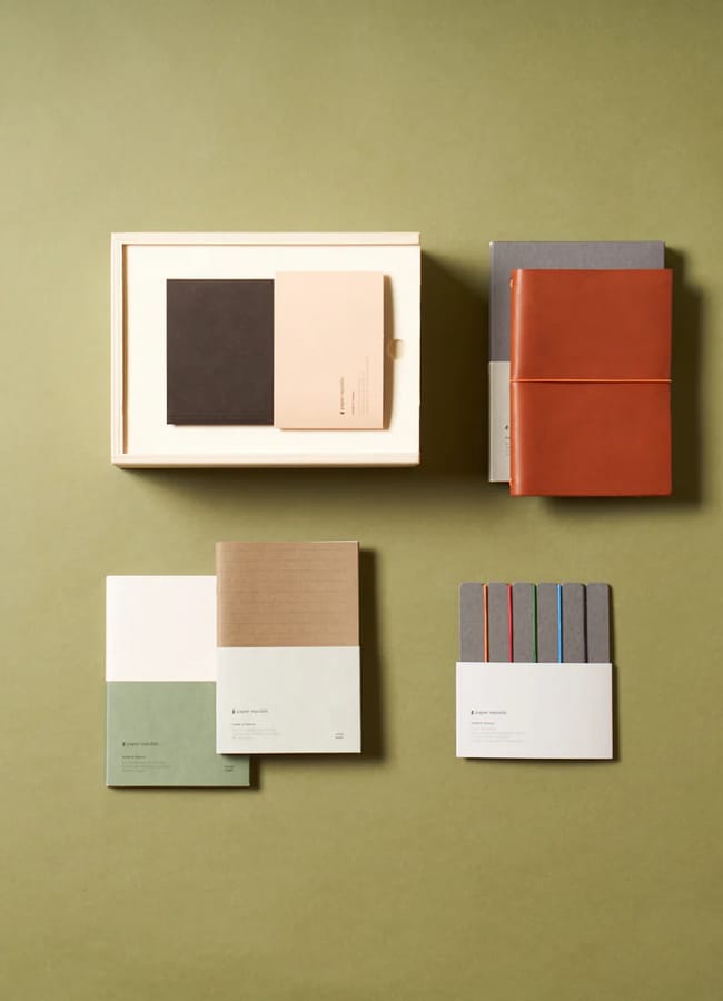 A flat lay of neatly arranged stationery items, including 2026 diaries with various coloured covers, a boxed set, and a set of slim notebooks, all on an olive-green background.