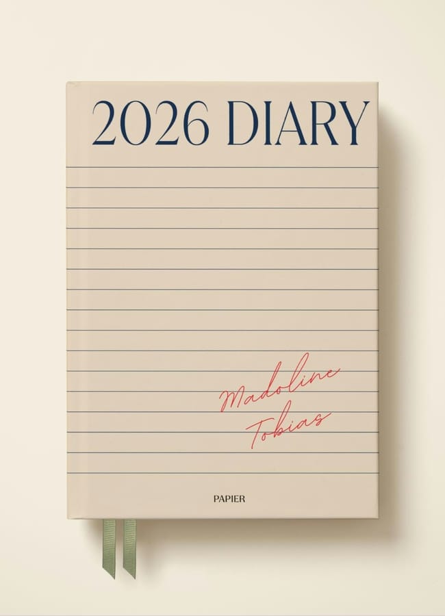 A beige 2026 diary with horizontal lines on the cover, 2026 DIARY in large blue text at the top, and Madeline Tobias written in red cursive. Ideal for fans of 2026 diaries, it features two green ribbons extending from the bottom.