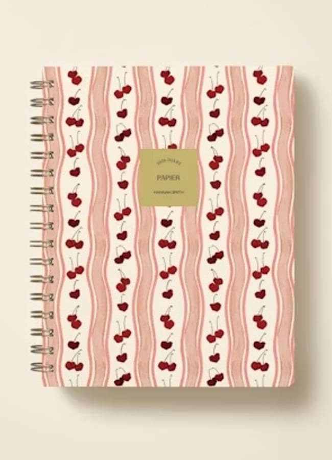 A spiral-bound notebook with a cream cover featuring wavy pink stripes and rows of red cherries, plus a gold label in the centre that reads PAPER—perfect for jotting notes or using as one of your 2026 diaries.