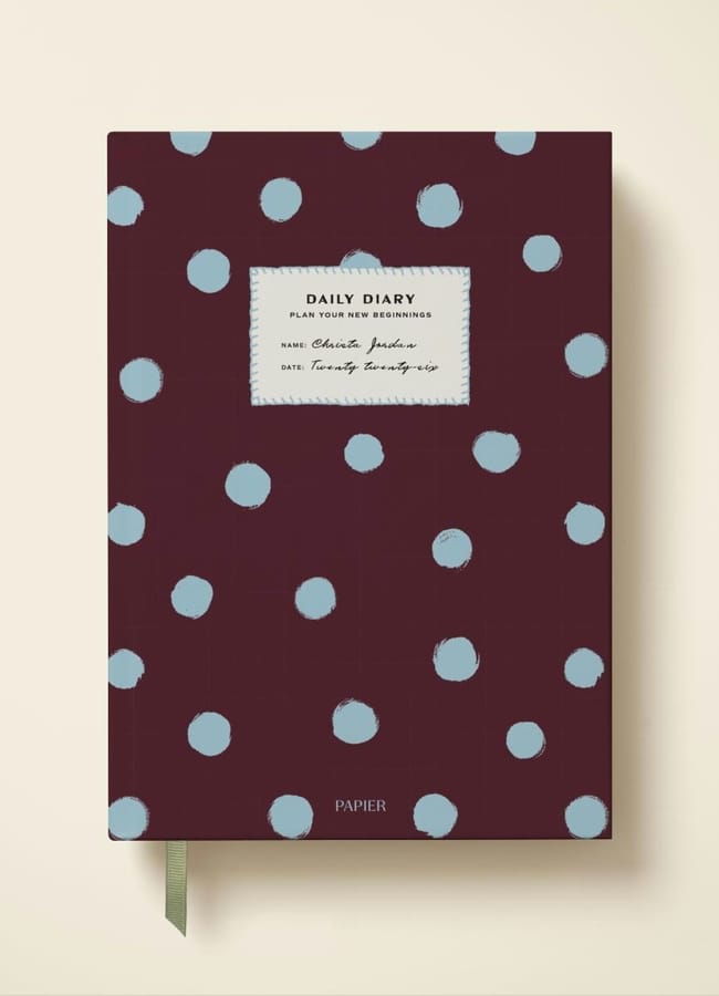 A maroon 2026 diary with light blue polka dots, a central white label, and a gold ribbon bookmark, placed on a light beige background.