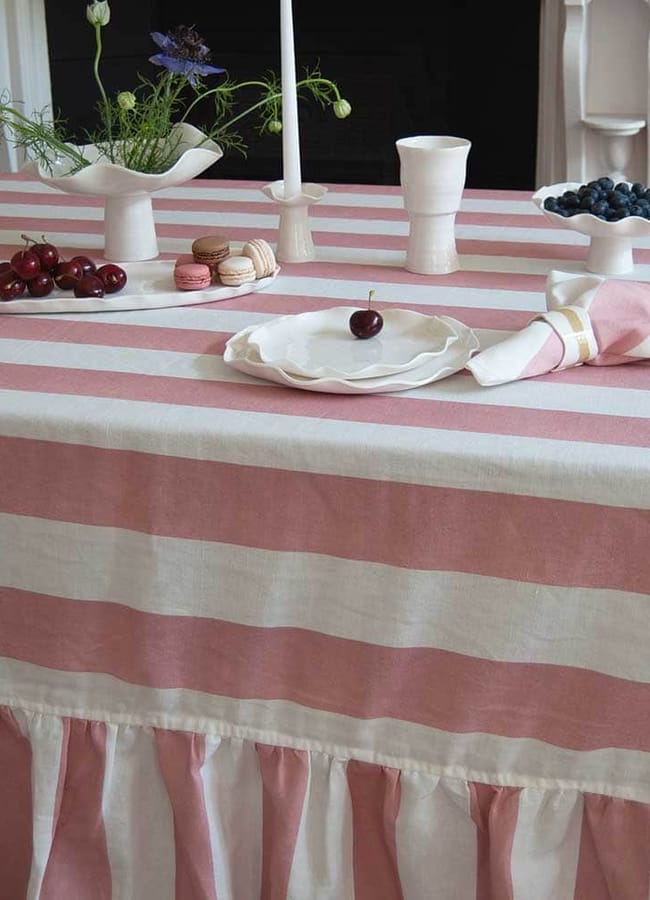 A table covered with a pink and white striped cloth, set for a charming Christmas tablescape with white crockery holding cherries, blueberries, and macarons, plus two white candlesticks and a small vase with flowers.