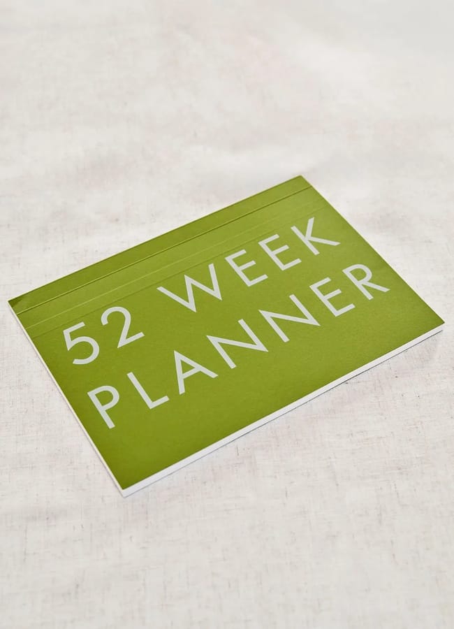 A green notebook with large white text on the cover that reads 52 WEEK PLANNER, placed on a light-coloured fabric surface—perfect for planning or alongside your 2026 diaries.