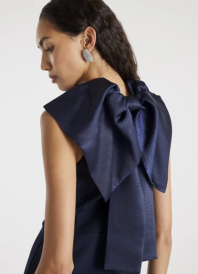 A woman with long dark hair wears a sleeveless navy dress, showcasing bow fashion with a large, shiny bow on the back and sparkly earrings. She stands facing away, looking down over her shoulder against a plain background.
