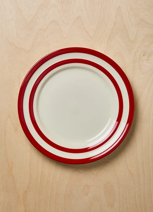 A round ceramic plate with two bold red rings near the edge, inspired by the Unexpected Red Theory, sits empty and centred on a light wood surface—a striking accent for contemporary interior design.
