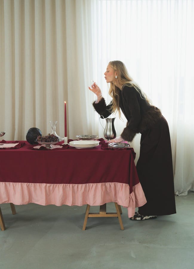 A woman with long blonde hair leans over a table with a red and pink tablecloth, embodying the Unexpected Red Theory in interior design as she lights a tall red candle. The table is laid with dishes, a jug, food, and sheer curtains in the background.