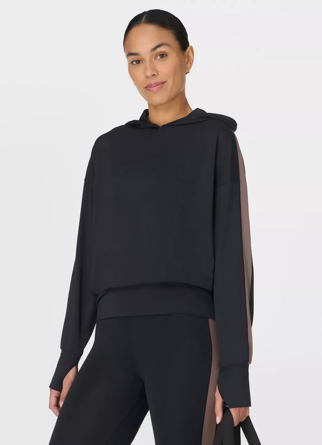 A woman wears the latest activewear: a loose black hoodie with brown stripes on the sleeves and matching black leggings with brown side panels, standing against a plain white background and holding a black bag.
