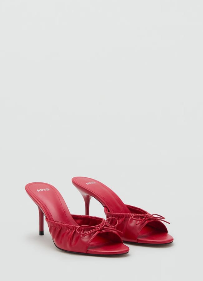 A pair of red high-heeled mules with open toes, thin heels, ruched detailing, and small bows on the front showcase timeless bow fashion on a plain light background.