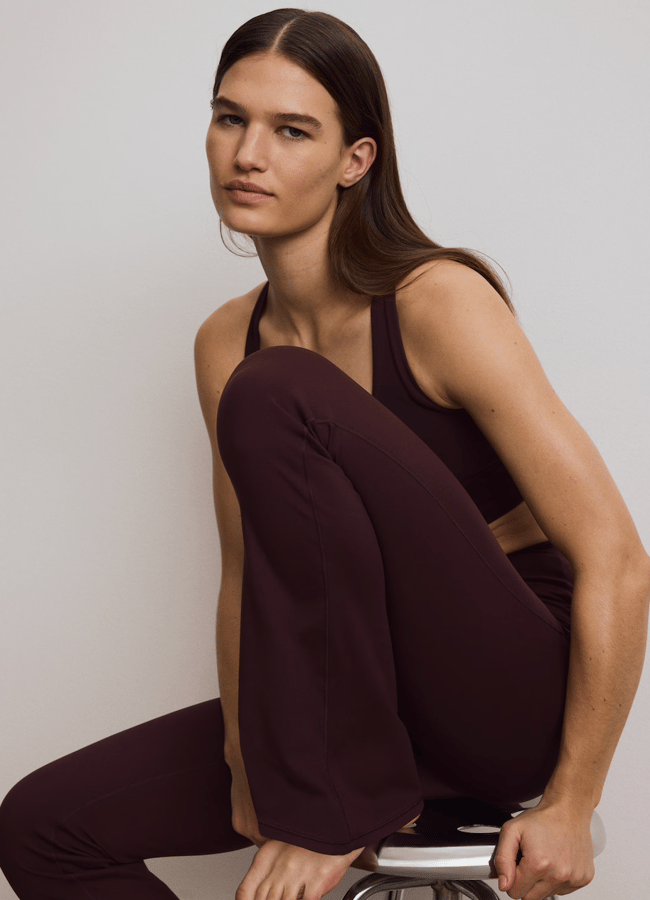 A person with long brown hair, wearing the latest sportswear—a dark brown sleeveless top and matching trousers—sits barefoot on a stool with one knee up, looking calmly at the camera against a plain light background.