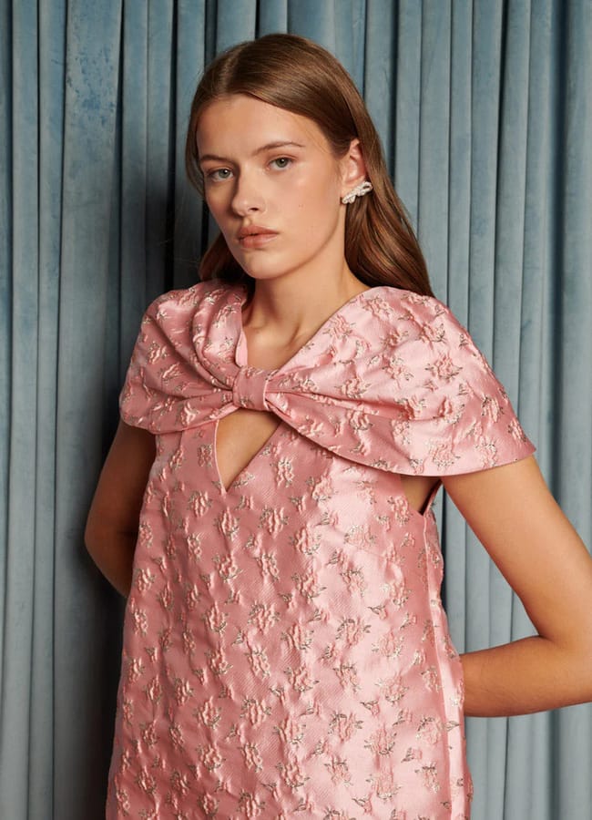 A woman stands in front of blue curtains wearing a textured pink dress, showcasing bow fashion with a large bow on the shoulders. She has straight, light brown hair and a neutral facial expression.