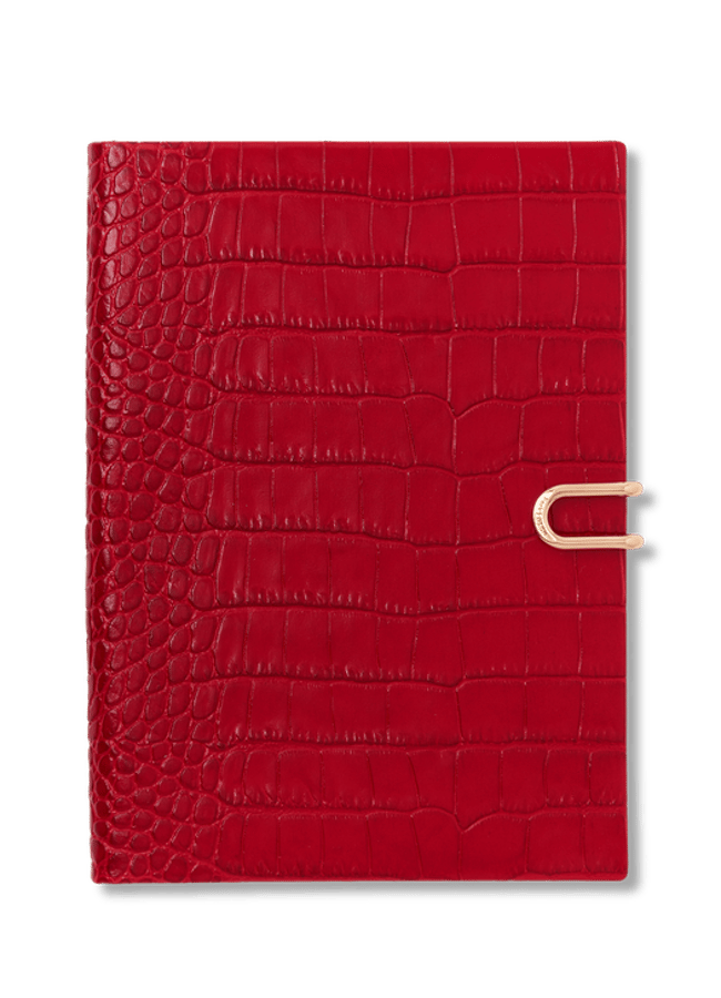 A square red notebook with a crocodile texture and a gold clasp on the right side, reminiscent of elegant 2026 diaries, set against a plain black background.