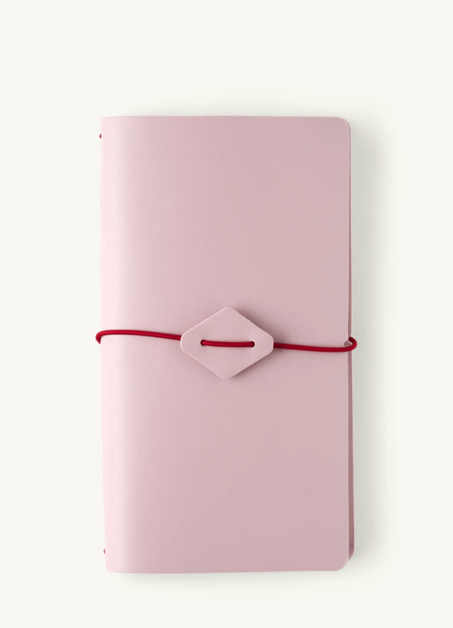 A closed, light pink 2026 diaries notebook with rounded corners, secured by a red elastic band threaded through a diamond-shaped clasp, placed on a plain white background.