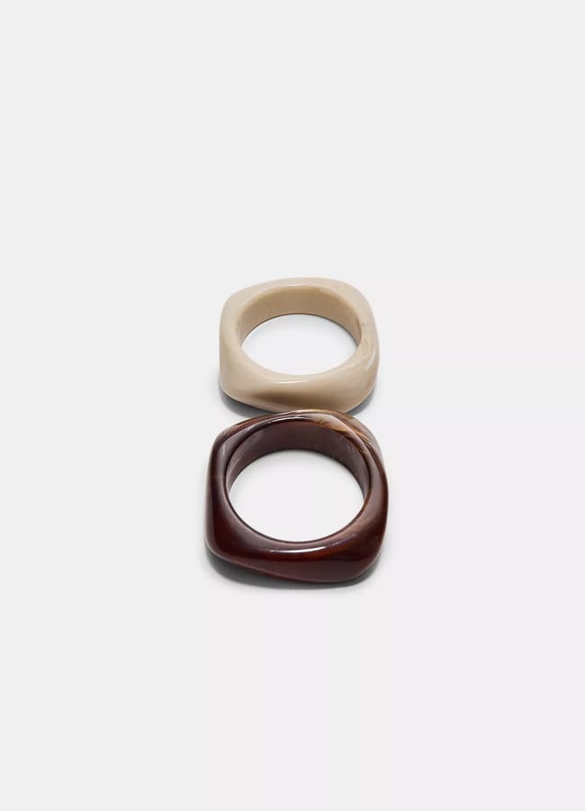 Two square-shaped resin rings, one beige and one dark brown reminiscent of classic brown trousers, are placed on a plain white background, arranged vertically with the beige ring above the brown ring.