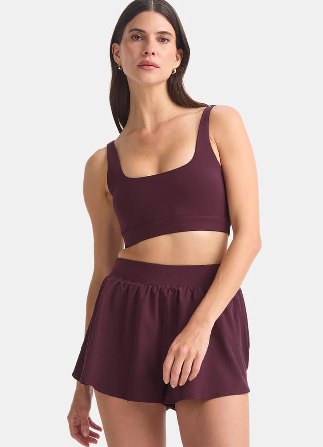 A woman wearing the latest activewear—a matching dark purple sports bra and shorts set—stands against a light grey background, looking forward with one hand by her side and the other holding her shorts.