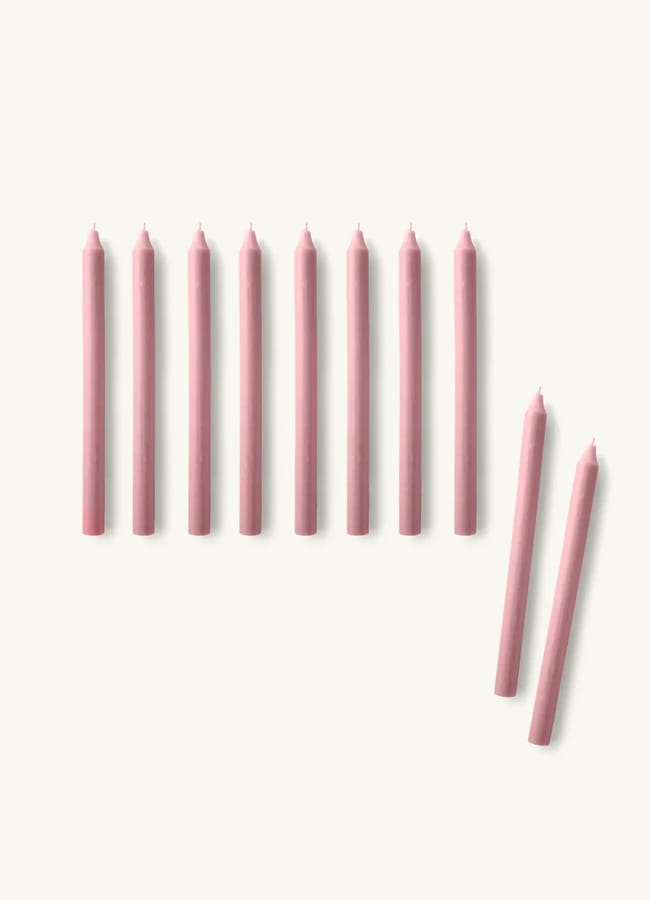 Ten pink taper candles are arranged upright in a row on a light surface, perfect for a Christmas tablescape. Two additional pink candles lie diagonally to the right of the group, separated from the row.