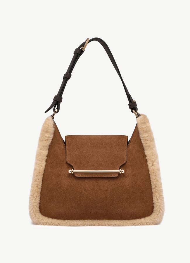 Brown suede handbag with a top handle, flap fastening, and light beige shearling trim. This chic fashion accessory features a gold bar accent on the front and an adjustable dark brown leather strap—perfect for pairing with trendy trousers or brown trousers.